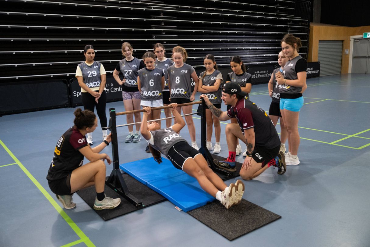 Young girl doing a pull up on bar, with group of female athletes around her, alongside YouFor2032 Talent Advisors.