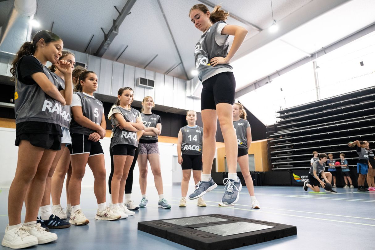Group of young female athletes watch another female athlete jump during a YouFor2032 testing day event.